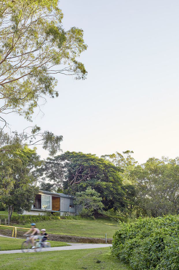 A compact modern single-storey dwelling nestled among mature trees in a lush landscaped setting. The house has a low horizontal profile, extensive glazing, dark metal or grey cladding, and warm timber wall accents. A paved path and blurred cyclists occupy the foreground, while dense greenery and a broad lawn surround the building.