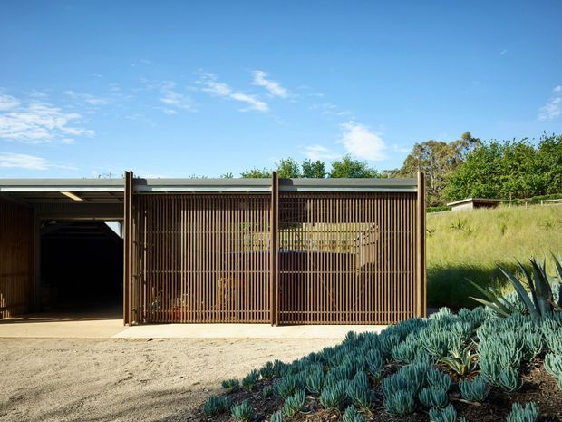 Exterior view of a low, contemporary structure set into a grassy landscaped site. The facade combines a flat, shallow roof with dark timber vertical screening and exposed structural posts, enclosing a wide open vehicle or storage bay on the left and a screened glazed or open area on the right. Gravel paving and sculptural blue-green succulent planting occupy the foreground, with trees and a sloping lawn behind.