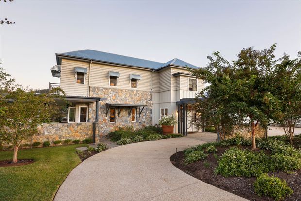 Exterior view of a substantial two-storey residence with a blue hipped/gabled roof, pale horizontal weatherboard cladding, and extensive irregular natural-stone walling around the lower level. The home features multiple rectangular windows, a small curved balcony, covered entry, and a broad curved concrete driveway or walkway through landscaped garden beds and lawn.