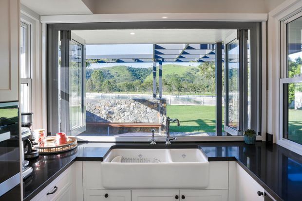 Bright, compact kitchen centered on a deep white farmhouse/apron-front sink beneath a large window opening. The room has white fixed cabinetry with dark hardware, glossy black countertops, a chrome gooseneck tap, white-framed glazing, and a broad outlook to a lawn, stone retaining wall, and green hillside. A retractable or roller blind is fitted above the main opening, and a covered outdoor structure is visible beyond.