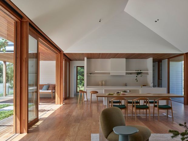 A bright, open-plan contemporary kitchen and dining area with extensive timber-framed glazing, warm timber flooring, and a timber-lined ceiling. The kitchen features minimalist white built-in cabinetry, open shelving, and a long island, while a timber dining table with green upholstered chairs occupies the foreground. Large windows and sliding doors connect the interior to a landscaped outdoor area, and a separate lounge or sitting area is visible through the glazing.