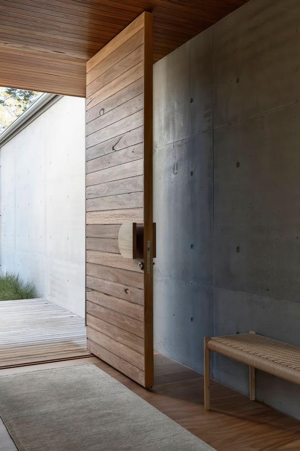 A minimalist residential entryway featuring a tall pivot door clad in horizontal natural timber boards. The entry has a warm timber-lined ceiling, dark timber flooring, a large neutral rug, and a simple woven bench. Raw grey concrete wall panels provide a contrasting industrial backdrop, while an open threshold leads to a timber deck and planted exterior area.