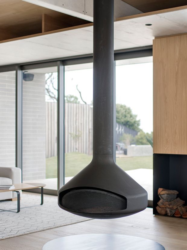 A contemporary open-plan living area featuring a suspended matte-black freestanding fireplace centered in front of full-height glazing. Pale timber cabinetry and ceiling joinery, light concrete or plasterboard surfaces, dark window frames, and a built-in firewood niche create a clean, restrained interior. A partial lounge setting and light rug are visible at left, with a leafy suburban garden beyond the glazing.