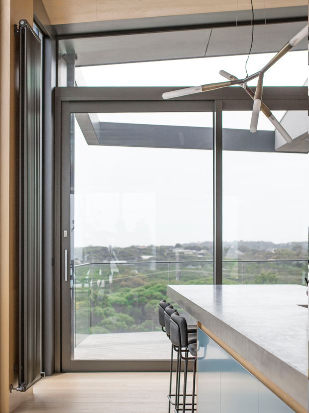 A contemporary kitchen interior with a long pale stone island and black bar stools positioned beside full-height glazed doors. The room features dark aluminium window and door frames, a timber ceiling and floor, a slim vertical radiator, and a sculptural linear pendant light. Beyond the glazing is a balcony with a glass balustrade and a leafy suburban outlook.