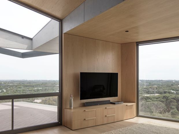 Contemporary living room with floor-to-ceiling corner glazing overlooking a leafy landscape, featuring a built-in light timber media wall and low television cabinet, timber ceiling, concrete accent panels, and a large deck visible outside.