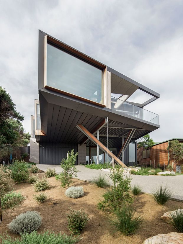 Exterior view of a contemporary multi-level residence on a landscaped site. The house features dramatic cantilevered upper volumes, extensive glazing, dark metal cladding, exposed structural steel and timber elements, a projecting balcony with clear glass balustrade, and a broad flat roof or terrace canopy. The front garden uses drought-tolerant ornamental grasses, shrubs, gravel and exposed soil.