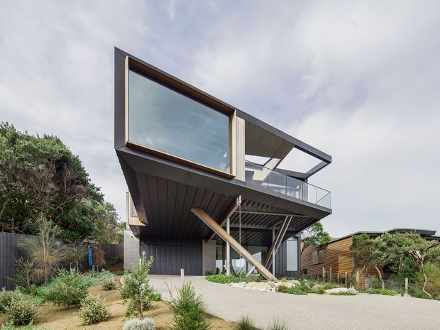 Exterior view of a contemporary elevated coastal-style residence with a dramatic cantilevered upper level, expansive dark-framed glazing, an angular flat roof, and a glazed balcony. The lower level is partially open with slender structural supports and a diagonal brace. Weathered timber cladding appears on the neighbouring structure, while the property is surrounded by drought-tolerant landscaping, gravel, native shrubs, and coastal vegetation.