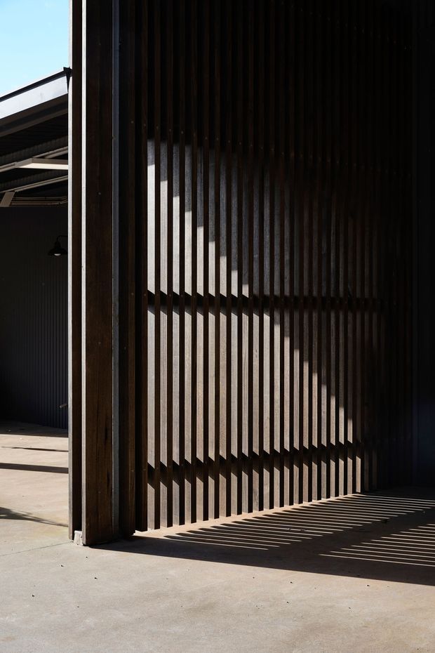 A dramatic architectural detail of a dark, industrial-style exterior or semi-enclosed structure. Tall vertical timber or dark metal slats form a screening wall, casting strong linear shadows across a smooth concrete floor and wall. Heavy structural posts, corrugated dark cladding, exposed roof framing, and a simple hanging light are visible, with bright sunlight entering from the left.