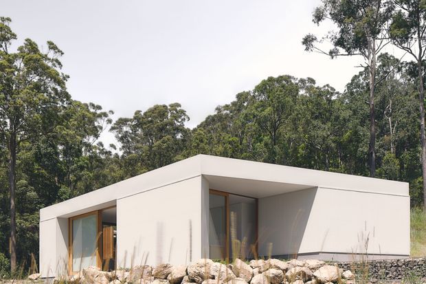A minimalist single-storey flat-roof house set within a wooded natural landscape. The building has crisp white rendered walls, deep roof overhangs and recessed openings, with warm timber-framed glazing and a low stone retaining wall in the foreground. Native trees and ornamental grasses surround the compact modern dwelling.