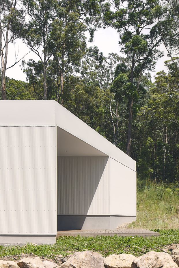 Exterior view of a minimalist, angular building set within dense eucalyptus bushland. The pale corrugated wall cladding and sharply sloping roof or canopy create a strong geometric facade, with a small timber deck and rocky landscaped foreground.