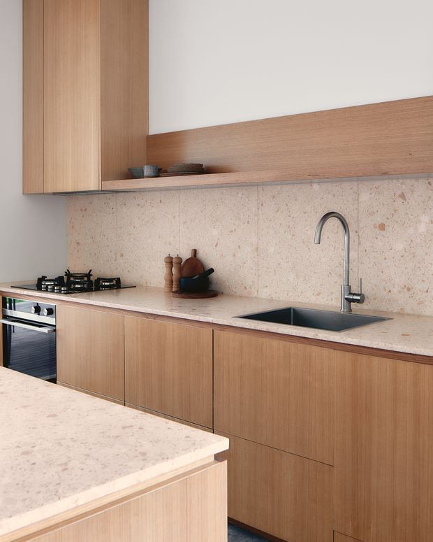 A refined contemporary kitchen with full-height and lower timber-look cabinetry, a pale pink-beige stone benchtop and matching slab splashback, integrated dark sink, curved chrome tap, gas cooktop and built-in oven. A long open timber shelf adds display space above the splashback, while a foreground island reinforces the functional kitchen layout. The restrained palette and clean lines create a warm, calm, minimalist feel.