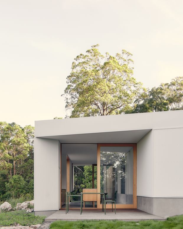 A minimalist single-storey modern house set within a leafy natural landscape. The facade features a crisp flat-roofed white volume, a recessed entry or outdoor sitting area, large timber-framed glazed doors, vertical ribbed wall cladding, and a grey lower wall base. A small outdoor dining setting sits on a simple paved platform, with mature trees and grasses surrounding the building.