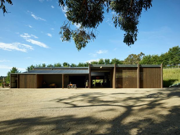 A low, single-storey modern building viewed from a broad gravel forecourt. The facade is composed of dark timber-clad volumes, open bays, vertical timber screening, and a shallow corrugated metal roof, with a covered central entry or passage. Mature trees and grassy planting surround the structure.