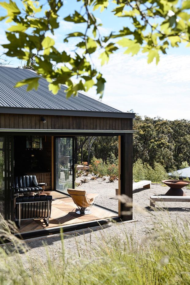 Exterior view of a compact, single-storey timber-clad dwelling with a dark corrugated metal pitched roof and expansive sliding glass doors opening onto a timber deck. The deck and interior lounge are visible through the glazing, while a gravel landscape with sculptural outdoor fire bowl and built-in seating surrounds the house. Mature vegetation and overhanging foliage frame the scene in bright daylight.