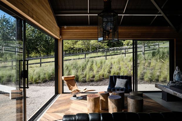 A dark, timber-lined living room with extensive floor-to-ceiling glazing overlooking a landscaped sloping garden. The room features warm herringbone timber flooring, black-framed glass doors and windows, a black leather sofa, sculptural log stools, a deep blue lounge chair, and a large hanging lantern. The space has a strong indoor-outdoor connection and a contemporary cabin-like character.