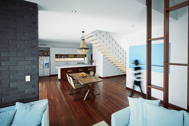 Bright open-plan contemporary interior combining a living area, dining space and kitchen. The room features dark timber flooring, a black textured masonry feature wall, white cabinetry, a timber dining table, pale blue sofas, and a distinctive white stair with open timber treads and circular cut-out detailing. Dark timber-framed glazing or screens define the right side of the space.