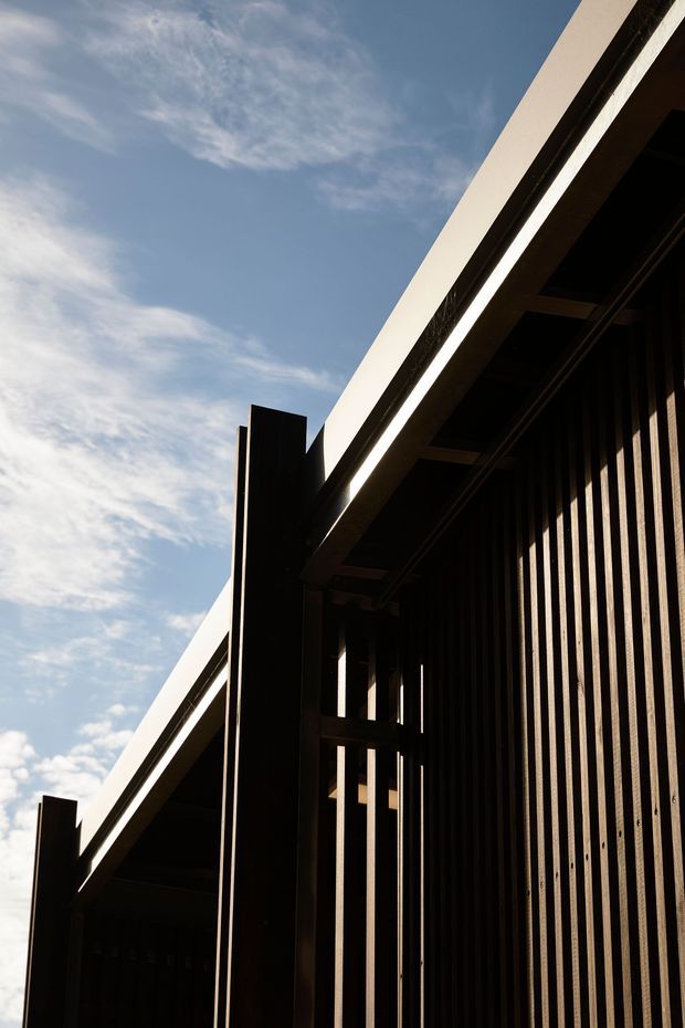 A dramatic upward-looking close-up of a contemporary building facade against a blue sky. The composition highlights dark vertical screening or cladding, deep structural fins, and a sharply projecting light-coloured roof edge or canopy, with strong sunlight creating high-contrast shadows.