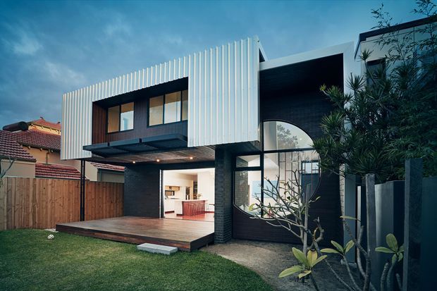 A contemporary two-storey residential house with a bold geometric facade. The upper level features vertical white cladding and dark-framed windows, while the ground floor combines dark brickwork, expansive glazing, and a projecting covered deck. A warm-lit kitchen is visible through the open rear-facing area, with landscaped garden elements and neighbouring homes surrounding the property.