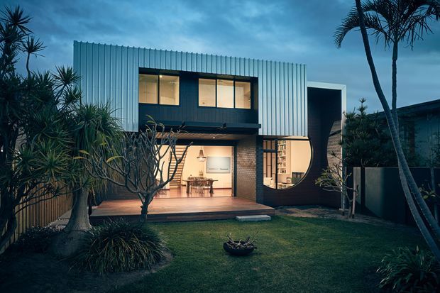 Exterior dusk view of a contemporary two-storey house surrounded by dense tropical and subtropical landscaping. The home has a corrugated light-coloured upper volume, dark cladding, exposed brickwork, large illuminated windows, and a covered timber deck opening toward a lawn. A distinctive large circular opening frames the ground-floor living area.