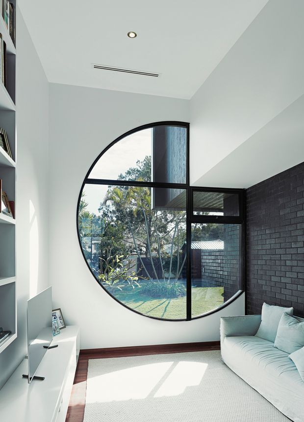 Bright contemporary living room with a pale upholstered sofa, white built-in shelving and low media cabinetry, dark timber floor trim, and a striking large circular black-framed window overlooking a landscaped courtyard. The room has white walls and ceiling, recessed lighting, a linear air vent, and a dark exposed-brick feature wall.