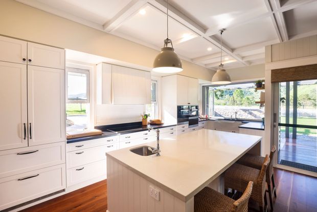 Bright, spacious kitchen with white shaker-style cabinetry, a large central island with integrated sink and seating, dark benchtops along the perimeter, and warm timber flooring. The room features exposed coffered ceiling beams, pendant lighting, expansive windows, and glazed doors opening toward an outdoor deck or garden.