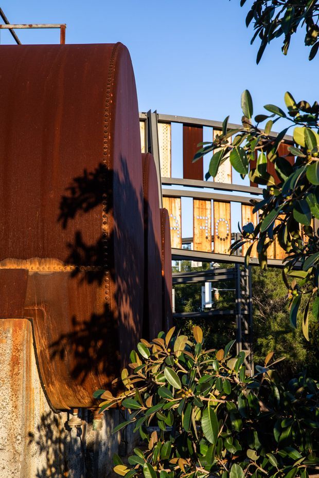 Exterior architectural detail featuring a large weathered rusted steel sculptural panel or tank-like facade element in the foreground, with a modern multi-level building behind it. The building has dark steel framing, extensive glazing, and warm timber or rust-toned infill panels, partially screened by dense leafy landscaping. Bright clear daylight emphasizes the contrast between the rusty metal, timber tones, and dark structural framing.