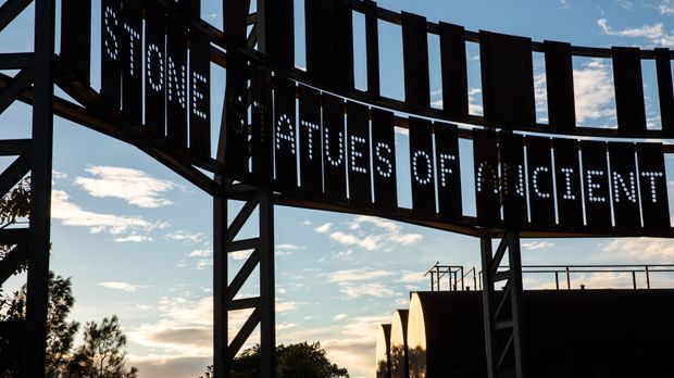Exterior architectural detail showing a large curved black metal sign structure with perforated lettering reading “Home of the Statues of Ancient…”. The sign is supported by exposed steel framing and set against an open sky, with part of a dark building visible behind it.