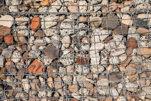 Close-up view of a gabion-style wall or structural cage filled with irregular natural stones and fragments in varied grey, beige, brown, and terracotta tones. The stones are held within a visible galvanized metal wire mesh grid, creating a strongly textured building-material surface.