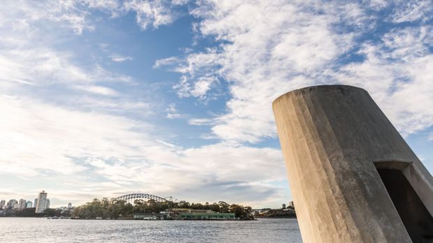 Exterior architectural photograph showing a large angled, weathered concrete structural element in the foreground beside a body of water. Across the water is an urban waterfront skyline with a distinctive steel arch bridge and tree-lined shore under a partly cloudy sky.