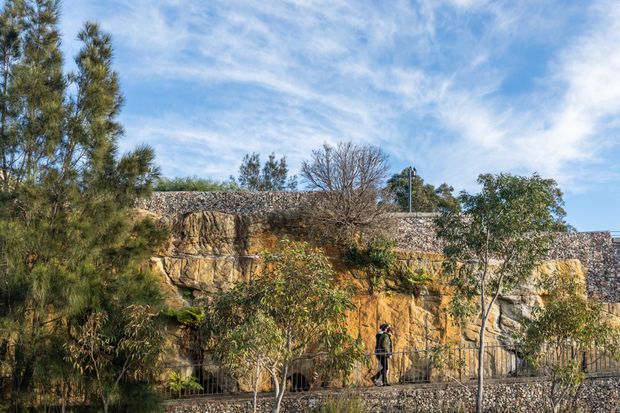 Exterior landscape view of a rocky, sandstone-like cliff and retaining wall integrated with a dry-stone or stone-clad structure. Native trees and shrubs frame the scene, with a pedestrian visible at the base. The image has a natural, rugged character under a bright blue sky.