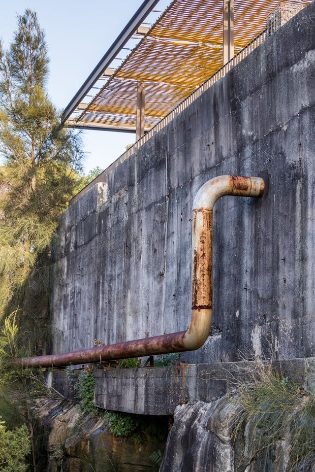 Exterior architectural detail showing a weathered concrete retaining wall with a large rusted metal pipe running across and upward along its face. Above the wall is a lightweight timber slatted pergola or screened canopy supported by steel posts. Native vegetation and rocky terrain surround the structure, creating an industrial, utilitarian landscape setting.