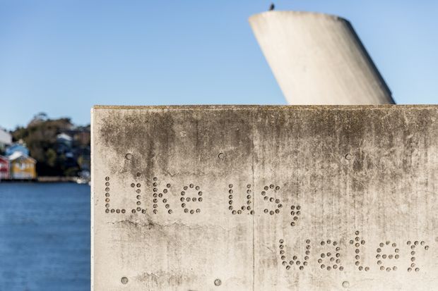 Close-up exterior view of a weathered concrete wall beside water, with perforated lettering spelling part of a place or building name. A large pale sculptural or structural concrete element rises behind the wall, with a distant waterfront setting visible to the left.