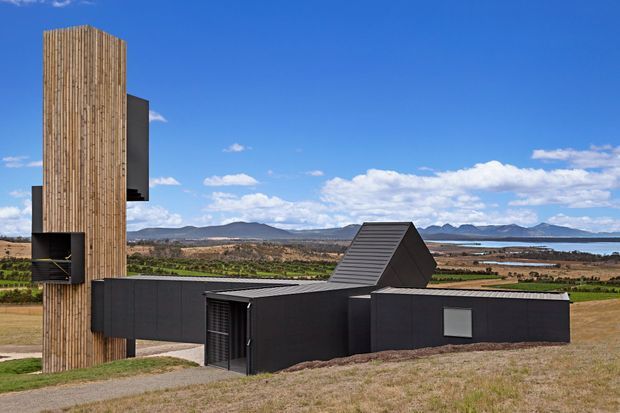 Exterior view of a striking contemporary rural building composed of dark cladding, a tall rounded vertical timber-clad tower, and angular pitched roof forms. The structure sits in an open agricultural landscape with distant mountains and a body of water under a bright blue sky.