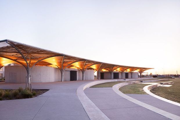 A wide exterior architectural view of a large contemporary pavilion or commercial complex at dusk. The long low structure has a sweeping translucent roof canopy supported by repeated branching steel columns, with warm concealed lighting beneath the canopy. Curved concrete pedestrian paths and landscaped open areas lead toward the building, which includes enclosed corrugated-metal or ribbed wall volumes and dark openings.