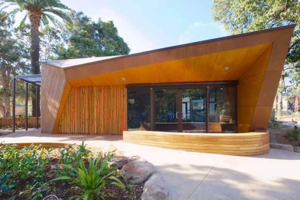 Exterior view of a distinctive low-slung timber-clad building with a strongly angled, folded roof and projecting canopy. Warm wood cladding continues across the soffit and facade, with a vertical timber feature wall beside dark-framed glazed doors. A curved built-in timber bench wraps the front, opening onto a paved forecourt and landscaped garden with mature palms.
