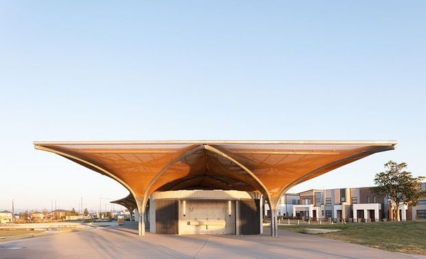 Exterior architectural view of a large sculptural tensile canopy forming a covered entry or gathering space in front of a contemporary building complex. The canopy has a broad, shallow roof with orange-brown fabric or panel surfaces, curved branching supports, and a central vaulted form. Low-rise modern buildings, paved forecourt, lawns, and sparse landscaping are visible beneath a clear sky.
