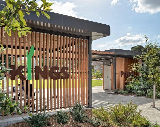 Contemporary low-rise building entrance with dark flat-roofed canopies, vertical timber screening, and large brown signage reading “KINGS” and partially visible “PA…”. A paved walkway leads between landscaped garden beds and lawn, with an open courtyard-like setting and bright daylight.