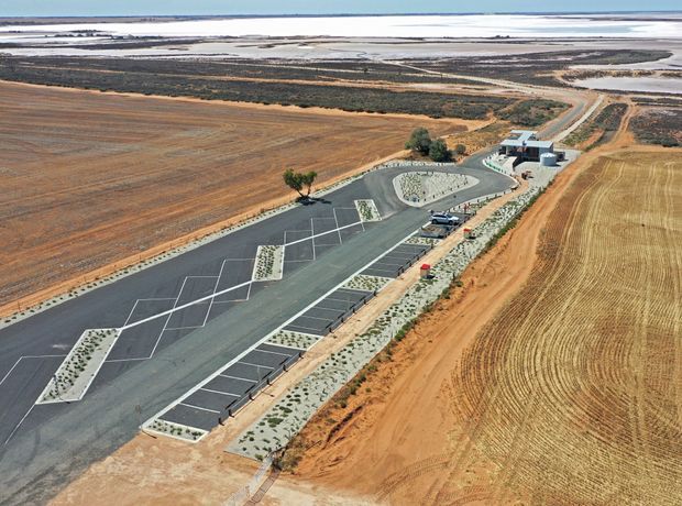 Aerial view of a newly constructed paved facility set within expansive agricultural fields beside a broad tidal or saltwater landscape. The site includes marked parking or hardstand areas, landscaped planting beds, access roads, and a small modern flat-roof building near the upper right.