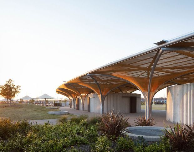 Exterior architectural view of a contemporary pavilion or canopy structure with sweeping curved roof forms, exposed steel supports, and corrugated metal-clad enclosed volumes. The building sits within a landscaped public or commercial setting, with planting beds, paved paths, and additional canopy structures visible in the background. Warm late-day sunlight highlights the timber-toned underside of the canopy.