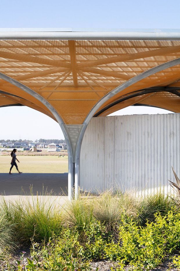 Exterior architectural view of a sculptural covered outdoor structure with branching steel supports and a warm orange woven or perforated canopy. The canopy shelters a paved pedestrian area beside a tall corrugated metal wall, with landscaped native-style grasses and shrubs in the foreground and open suburban grounds beyond.