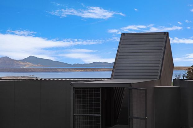 Exterior architectural view of a dark, highly geometric structure beside a large body of water. The building has a low horizontal roofline, a prominent steeply angled louvred or ribbed roof/wall element, dark metal cladding, and a secure grid-like metal gate. Mountains and blue sky form the distant background.