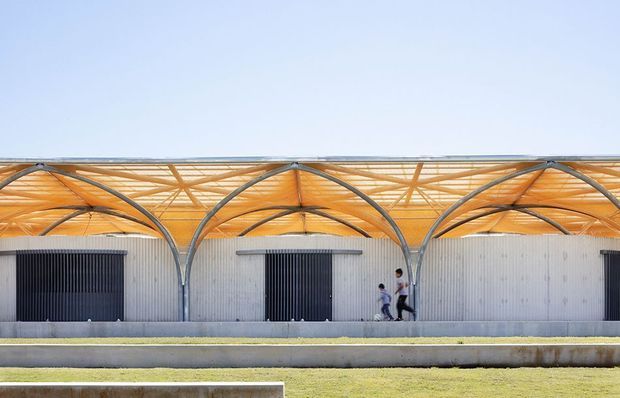 Exterior architectural view of a long, low industrial or commercial building with corrugated light-grey metal walls, large dark roller doors, and a repeated sculptural arched roof canopy. The canopy has exposed curved steel ribs and warm timber-toned roof lining, creating a rhythmic bays-and-arches facade. A paved forecourt and landscaped grass strips sit in front, with two people providing scale.