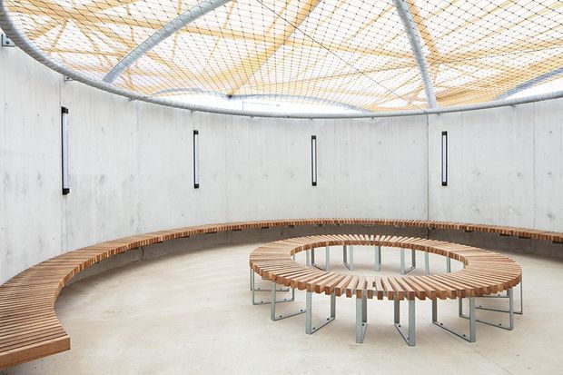A bright circular architectural interior with smooth exposed concrete walls, a curved perimeter bench, and a central circular timber-slat bench surrounding a glazed floor opening. Narrow vertical windows punctuate the walls, while a lightweight curved steel-and-mesh roof structure creates an airy patterned ceiling.
