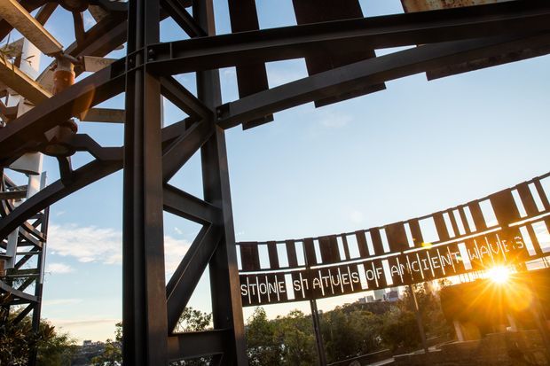 Exterior architectural detail of a steel-framed structure with prominent dark metal beams, diagonal bracing, and an elevated walkway or sign railing. The sun is low behind the structure, with blue sky and trees in the background. The image emphasizes industrial construction and exposed structural steel.