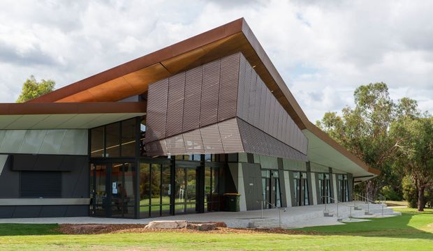 Exterior view of a contemporary single-storey building with a dramatic asymmetric folded roof, broad roof overhangs, dark framed floor-to-ceiling glazing, and a prominent suspended or projecting louvred facade screen. The building sits within landscaped grass and trees under a partly cloudy sky.