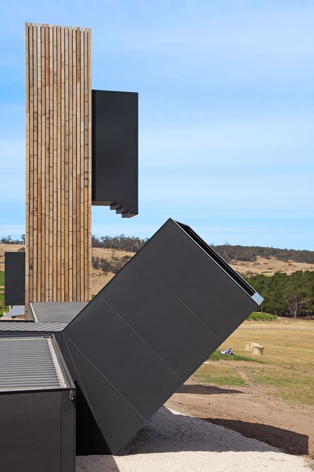 A striking modern building exterior in a rural landscape, composed of sharply angled black-clad geometric volumes and a tall vertical tower faced with closely spaced natural timber battens. Corrugated metal roofing and dark wall panels are visible in the foreground, with open grassland and low trees beyond under a clear blue sky.