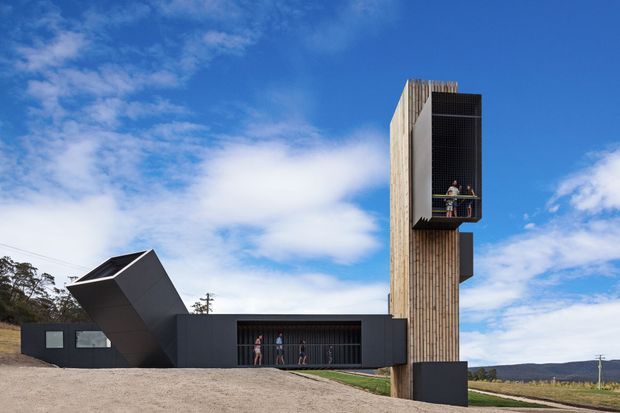 A striking contemporary architectural complex set in an open rural or semi-rural landscape. The composition features a tall vertical tower clad in light timber battens, with a dark recessed upper opening and balcony, connected to a low dark rectangular wing. A sharply angled, faceted dark volume projects from the opposite side. The building is photographed externally against a vivid blue sky, with people visible near the sheltered openings for scale.