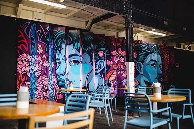 A colourful contemporary café or commercial dining space with round wooden tables, turquoise metal chairs, exposed ceiling services, and a large vivid floral and portrait mural covering the rear wall. The atmosphere is bold, artistic, and casual.