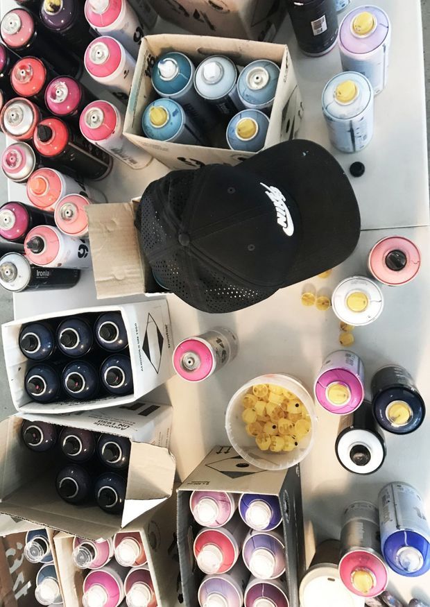 Top-down photograph of numerous colorful spray paint cans arranged in open cardboard boxes and scattered across a light work surface. A black Nike cap lies near the center, with a small bowl of yellow snack pieces nearby. The image depicts an art or workshop setup rather than an architectural space.