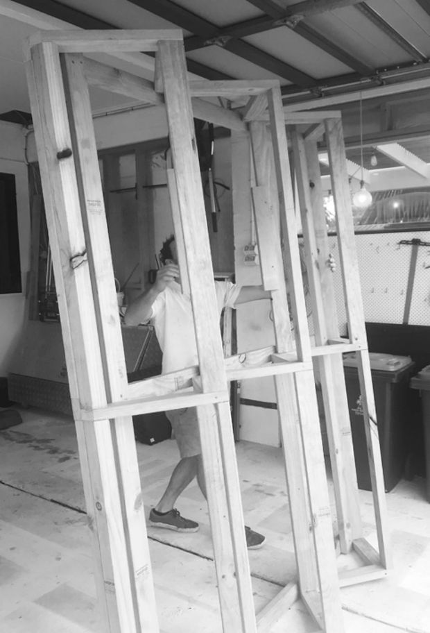 Black-and-white construction photograph showing an unfinished timber-framed partition or wall assembly inside a workshop or building site. Exposed light timber studs and horizontal bracing dominate the foreground, with an exposed metal roof structure overhead and construction materials and equipment in the background. A partially obscured worker is visible behind the frame.